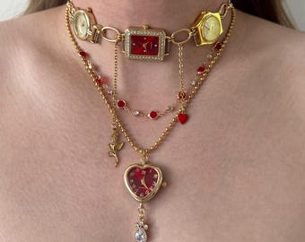 May include: A gold-toned necklace featuring multiple watch faces and decorative elements. The necklace includes a rectangular watch with a red face, heart-shaped watch pendant, and red and clear gem accents. The design incorporates a rose and a dangling teardrop-shaped gem.