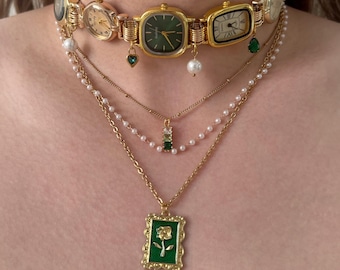 May include: A gold-toned choker necklace featuring multiple vintage watch faces. Below, layered necklaces include pearl strands and pendants with green gemstones and a green framed rose design.