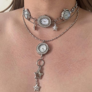 Op de afbeelding: Zilverkleurige ketting met meerdere horloges en sterbedels. De choker-stijl ketting heeft drie horloges aan de bovenkant, met een langere ketting met een bungelend horloge en sterhangers. De horloges hebben witte wijzerplaten en zilveren randen.