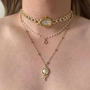 Op de afbeelding: Een gelaagde ketting set. De bovenste laag is een goudkleurige horlogehalsband, gevolgd door een parelketting met een sterhanger en een gouden ketting met een pareldruppel. De kettingen zijn ontworpen om samen gedragen te worden.