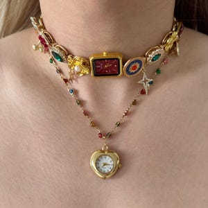 Può includere: Una collana dorata con un orologio rettangolare con quadrante rosso e un ciondolo a forma di cuore. La collana è impreziosita da pietre preziose colorate e ciondoli decorativi, creando un effetto a strati.