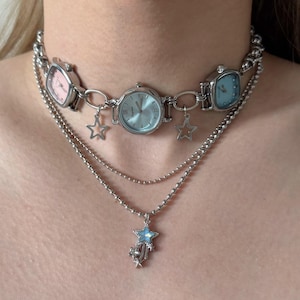 May include: A silver-toned necklace featuring a choker with three watch faces in pink and blue. The necklace has two additional strands with star charms. The watch faces have silver bezels and light-colored watch faces.