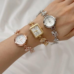 May include: Three elegant wristwatches with white faces and delicate bands. One watch has a rose gold finish, another is gold-toned, and the third is silver. Each watch features a unique face shape and decorative accents, including floral and gemstone details.