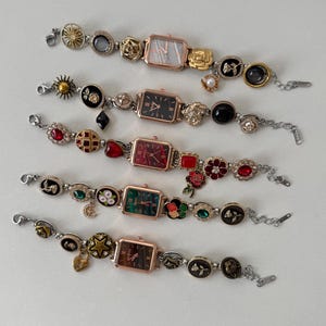 May include: Five gold-tone wristwatches with different coloured faces and decorative charms. The charms include flowers, stars, hearts, and other designs.