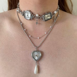 May include: A silver-toned necklace featuring multiple watch faces, pearls, and a heart-shaped pendant with a teardrop pearl. The necklace has a layered design with a choker-style top layer and a longer pendant.