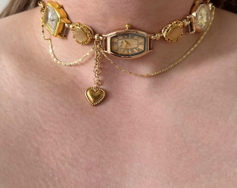 May include: Gold-toned watch choker necklace featuring multiple watch faces and heart pendant. The necklace has a delicate chain design with a central rectangular watch and smaller watch faces on each side. The heart pendant hangs from a chain.