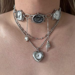 May include: A silver necklace with multiple watch faces. The choker-style band has three watch faces, and there are dangling chains with heart and hand charms. The watch faces display Roman numerals. The necklace is a unique piece of jewellery.