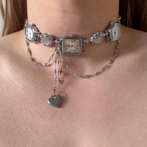 Könnte beinhalten: Ein silberfarbenes Choker-Halsband mit mehreren kleinen Zifferblättern und dekorativen Elementen. Die Halskette hat eine zentrale rechteckige Uhr mit kleineren runden Uhren auf jeder Seite. Eine Kette mit einem Herzanhänger hängt von der Mitte herab.