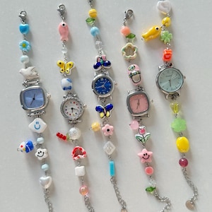 May include: Six colorful beaded watch bracelets with different charms, including butterflies, flowers, fish, and smiley faces. Each bracelet has a small, round watch face with silver hands.