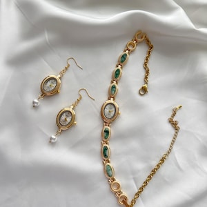 May include: A gold-toned watch bracelet and matching earrings set. The bracelet features oval-shaped watch faces and green stone accents. The earrings have a watch face design with a dangling pearl. The jewelry is displayed on a white, textured surface.