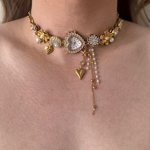 May include: Gold-toned choker necklace featuring a heart-shaped watch, floral accents, and dangling charms. The necklace includes pearls, stars, and a heart pendant. The watch face has white background with black numbers.