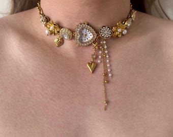 Może przedstawiać: Złoty naszyjnik typu choker z zegarkiem w kształcie serca, kwiatowymi akcentami i wiszącymi zawieszkami. Naszyjnik zawiera perły, gwiazdki i zawieszkę w kształcie serca. Tarcza zegarka ma białe tło z czarnymi cyframi.