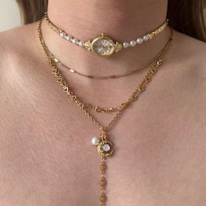 Puede incluir: Conjunto de collares dorados en capas. La capa superior es un choker con una esfera de reloj y detalles de perlas. La segunda capa es una cadena fina. La tercera capa presenta un colgante de media luna con una cadena en forma de corazón y una perla.