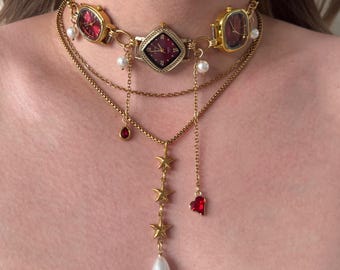 May include: Gold-coloured watch choker necklace with red watch faces and dangling charms. The necklace features watch faces, pearls, stars, and a heart pendant. The watch faces have a burgundy-coloured face.