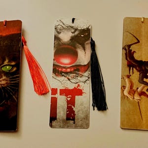 May include: Three bookmarks with different horror movie themes. The first bookmark features a close-up of a black cat with green eyes and sharp teeth. The second bookmark features a close-up of a clown's face with a red nose and the word "IT" in red. The third bookmark features a black figure with a scythe attacking a woman in a white dress.