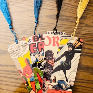May include: Four vintage comic book bookmarks with colorful tassels. The bookmarks feature images of superheroes and the text "THE HAND OF", "BLOR", "ER", and "B123".