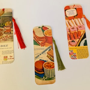 May include: Three vintage bookmarks with colorful images and text. The bookmarks feature advertisements for Hunt's Tomato Sauce, Campbell's Soup, and a brand of canned meat. Each bookmark has a tassel.