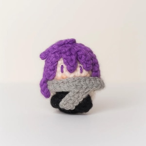 Könnte beinhalten: Handgemachte Häkelpuppe mit lila Haaren, grauem Schal und schwarzer Kleidung. Die Puppe hat einen hellen Hautton und ist vor einem schlichten weißen Hintergrund platziert. Die Merkmale der Puppe sind einfach, mit kleinen, genähten Augen.