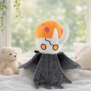 Può includere: Un peluche raffigurante un personaggio con una testa arancione a forma di fungo con macchie rosa, corna bianche e un viso bianco con occhi arancioni. Il personaggio ha un corpo grigio a forma di mantello ed è su una superficie bianca.