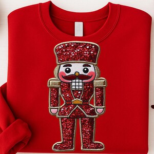May include: A red sweatshirt featuring a sequined nutcracker design. The nutcracker is detailed with gold accents and a red sequined uniform. The sweatshirt is a solid red color, perfect for the holiday season.