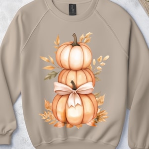 May include: A beige sweatshirt with a watercolour design of three stacked pumpkins and a bow, surrounded by autumn leaves. The pumpkins are orange and white, with a light beige bow. The leaves are in shades of orange and yellow.