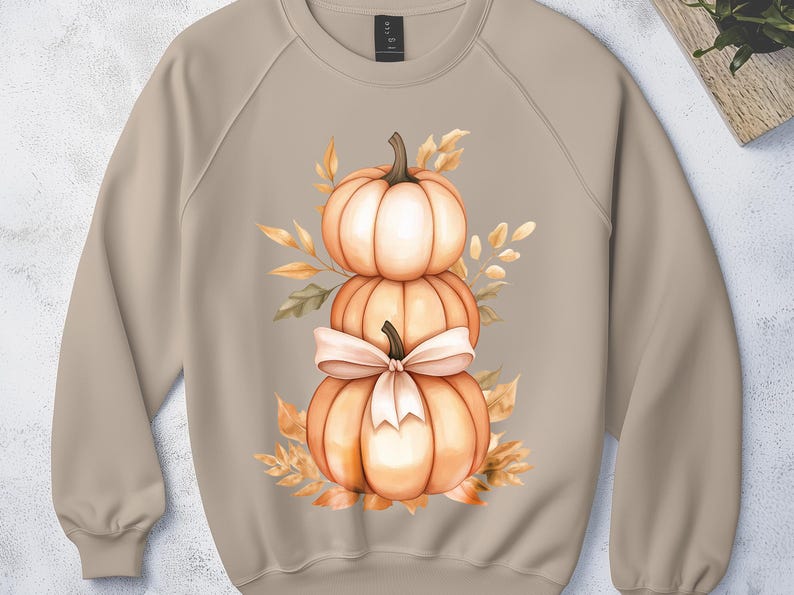 May include: A beige sweatshirt with a watercolour design of three stacked pumpkins and a bow, surrounded by autumn leaves. The pumpkins are orange and white, with a light beige bow. The leaves are in shades of orange and yellow.