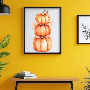May include: A framed watercolour painting of three stacked pumpkins tied with a ribbon. The artwork is displayed on a yellow wall, next to a framed fern print. A wooden bench with books and plants completes the scene.