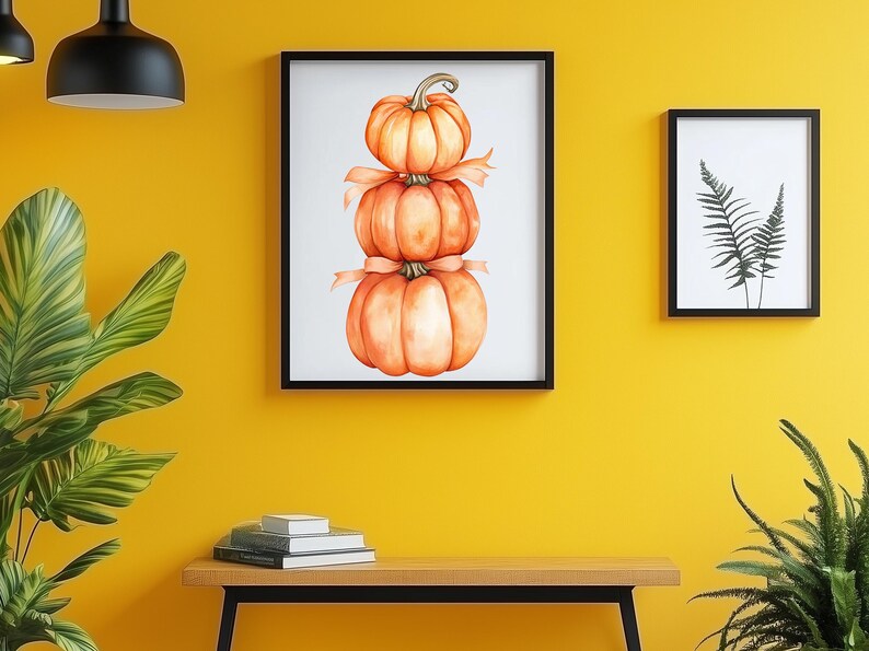 May include: A framed watercolour painting of three stacked pumpkins tied with a ribbon. The artwork is displayed on a yellow wall, next to a framed fern print. A wooden bench with books and plants completes the scene.