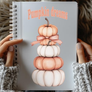 May include: A notebook with a spiral binding and a grey cover. It features a stack of pumpkins in orange and white, with a bow. The words "Pumpkin dreams" are written in orange script above the pumpkins.