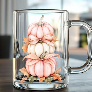 May include: A clear glass mug with a handle, featuring a stacked pumpkin design. The pumpkins are in shades of pink and white, adorned with bows and autumn leaves. The mug is set on a wooden surface.