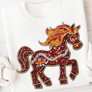 Op de afbeelding: Witte sweatshirt met een geborduurd paardenontwerp. Het paard is rood met een gouden manen en staart, en is versierd met kleurrijke, sprankelende accenten. Het ontwerp is gecentreerd op de voorkant van de sweatshirt.