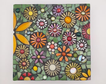 May include: A square mosaic art piece featuring a colorful floral design. The mosaic is made of small, colorful tiles arranged in a pattern of flowers and leaves. The background is a dark green color.