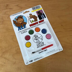 May include: A plastic paint set featuring the character ALF. The set includes six non-toxic watercolors, a paintbrush, and a custom ALF palette. The packaging is white with blue and red accents. The text on the packaging reads "ALF Paint Set" and "6 Non-Toxic Watercolors Paint Brush Custom ALF Palette".