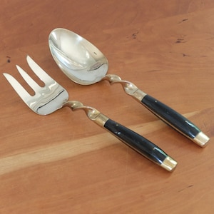 May include: A serving fork and spoon set. The utensils have silver-colored heads and twisted brass-colored stems. The handles are black with brass-colored accents. The set is displayed on a wooden surface.