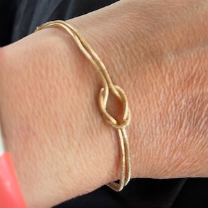 May include: A gold-coloured bracelet featuring a knot design. The bracelet is made of thin, textured metal strands. The knot is the focal point, with the bracelet wrapping around the wrist. The background is a dark fabric.