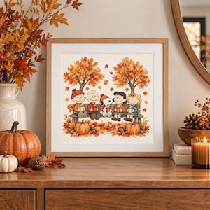 May include: A square framed wall art print featuring a watercolor illustration of the Peanuts characters sitting on a wooden bench under autumn trees. Charlie Brown, Snoopy, and friends hold mugs amidst falling orange leaves and pumpkins.