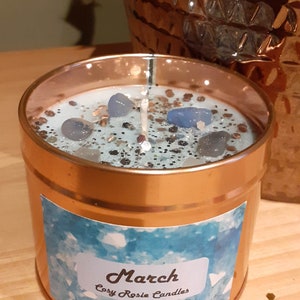 May include: A copper candle jar with a blue and silver glitter candle inside. The candle has blue crystal pieces embedded in it. The jar has a label that says "March Cosy Rosie Candles".