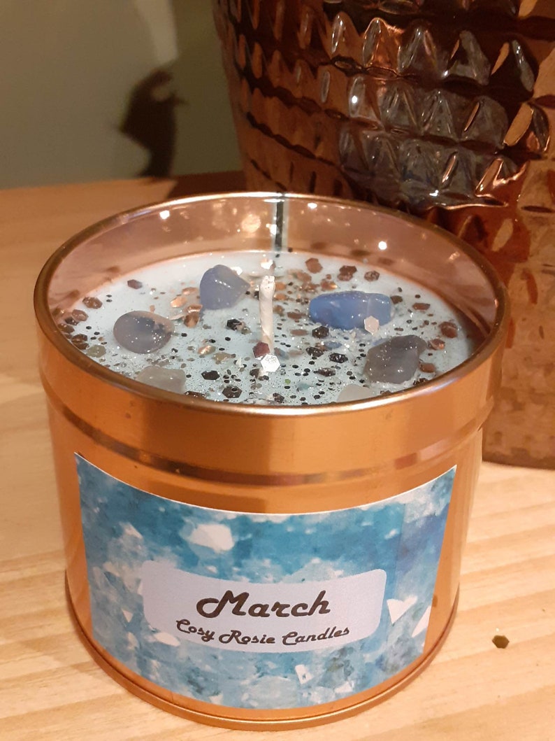 May include: A copper candle jar with a blue and silver glitter candle inside. The candle has blue crystal pieces embedded in it. The jar has a label that says "March Cosy Rosie Candles".