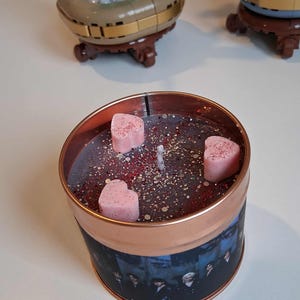 May include: A copper-coloured candle tin with a dark wax interior, speckled with glitter and small red accents. Three pink heart-shaped wax melts are placed on top. The tin's exterior features a printed image of a group of people.