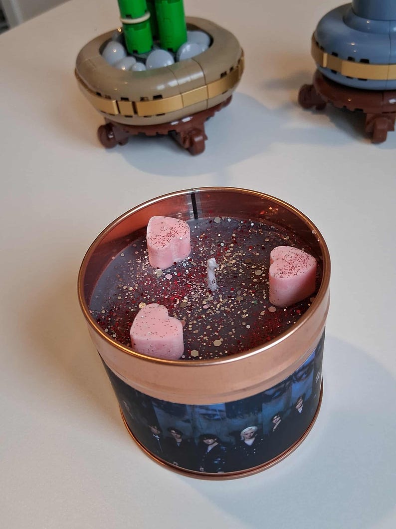 May include: A copper-coloured candle tin with a dark wax interior, speckled with glitter and small red accents. Three pink heart-shaped wax melts are placed on top. The tin's exterior features a printed image of a group of people.
