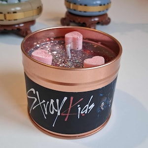 May include: A copper-coloured candle tin with a black label featuring the text "Stray Kids" in white and red. The candle contains pink heart-shaped wax melts and glitter. The candle is a decorative item.