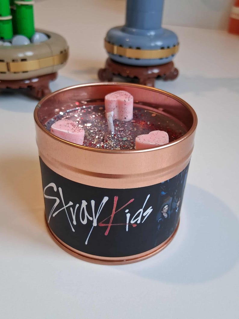 May include: A copper-coloured candle tin with a black label featuring the text "Stray Kids" in white and red. The candle contains pink heart-shaped wax melts and glitter. The candle is a decorative item.