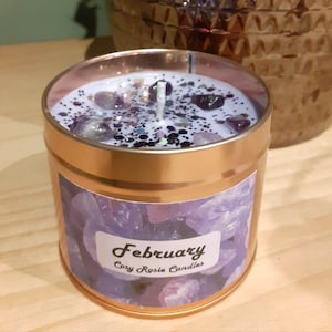 May include: A gold candle tin with a purple and white label that says "February" and "Cory Rosie Candles". The candle is topped with a white wax layer and purple and white glitter.