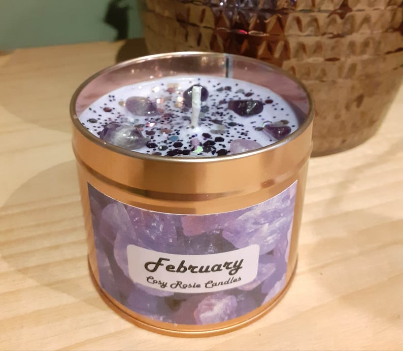 May include: A gold candle tin with a purple and white label that says "February" and "Cory Rosie Candles". The candle is topped with a white wax layer and purple and white glitter.