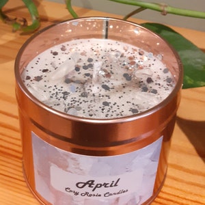May include: A copper-colored candle jar with a white label that says "April Cozy Rosie Candles". The candle is topped with a layer of white wax and silver glitter.