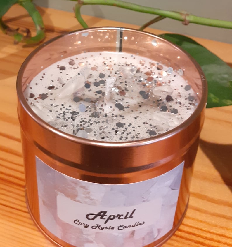 May include: A copper-colored candle jar with a white label that says "April Cozy Rosie Candles". The candle is topped with a layer of white wax and silver glitter.