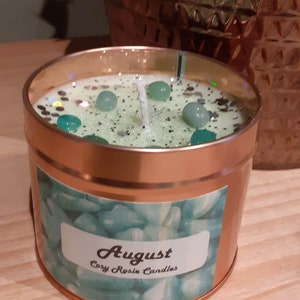 May include: A copper candle jar with a green and white label that says "August" and "Cozy Rosie Candles". The candle is topped with a white wick and green glitter.