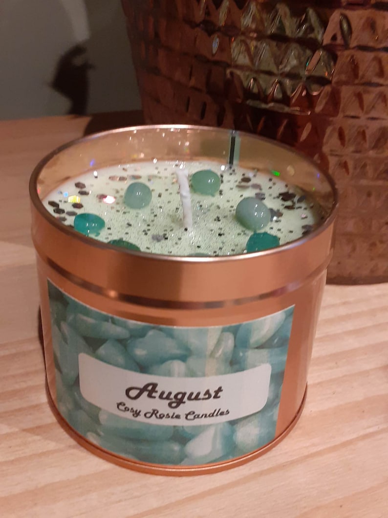 May include: A copper candle jar with a green and white label that says "August" and "Cozy Rosie Candles". The candle is topped with a white wick and green glitter.