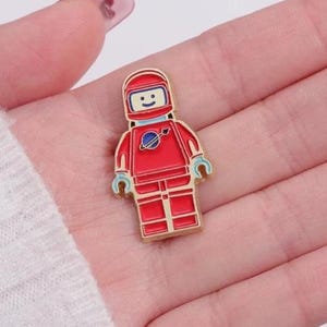 May include: A red astronaut pin badge with a smiling face on the helmet and a blue planet emblem. The pin has a gold border. The astronaut is wearing a red spacesuit with blue detailing.