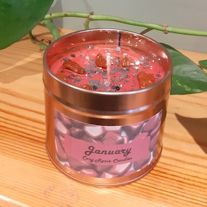 May include: A red candle in a copper tin with a silver glitter and brown crystal topping. The label reads "January Cozy Rosie Candles".